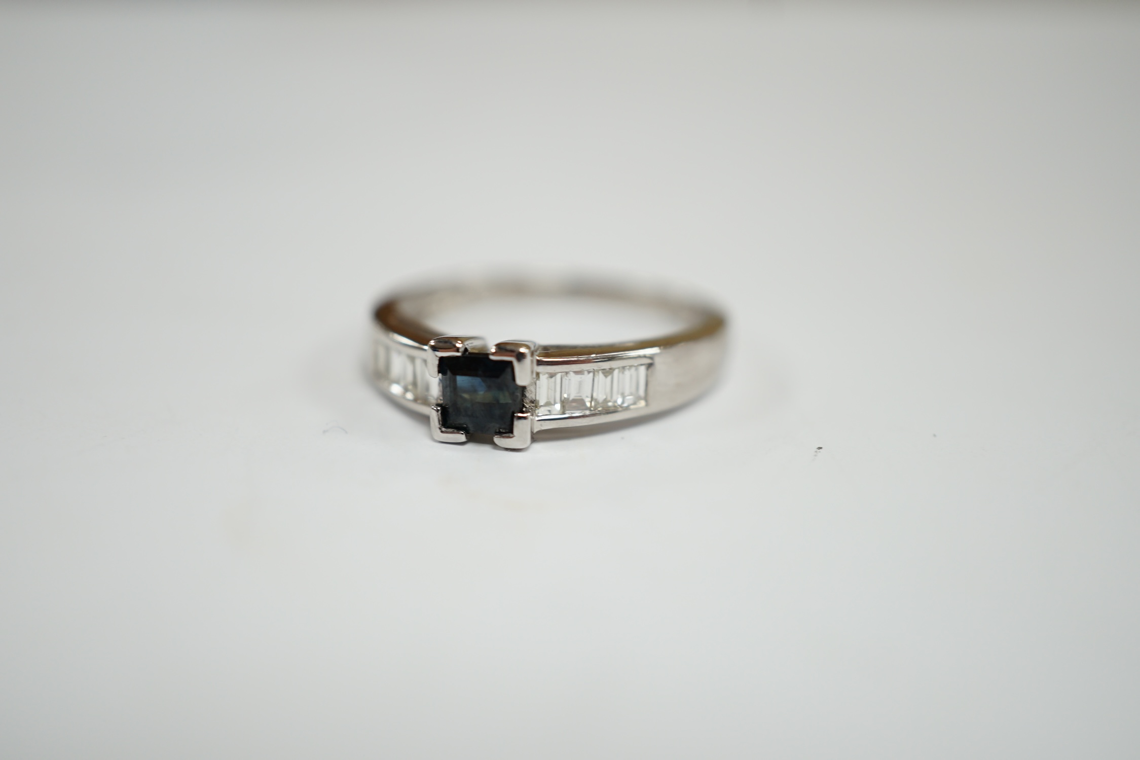 A modern 18ct white gold and single stone square cut sapphire set ring, with baguette cut diamond set shoulders, size N, gross weight 2.9 grams.